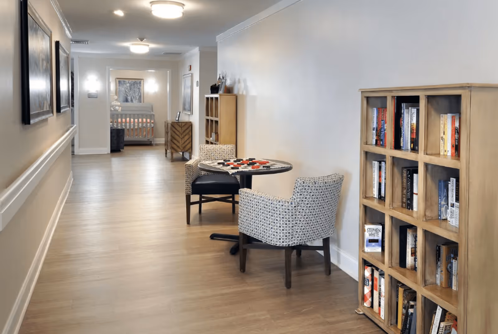 A well-lit hallway in a senior living facility with light wood flooring and neutral-colored walls. On the right side, there is a wooden bookshelf filled with books and a small round table with a checkerboard game set up, accompanied by two patterned armchairs. At the end of the hallway, a room with a bed and framed artwork is visible. The walls are decorated with framed pictures and wall sconces provide additional lighting.