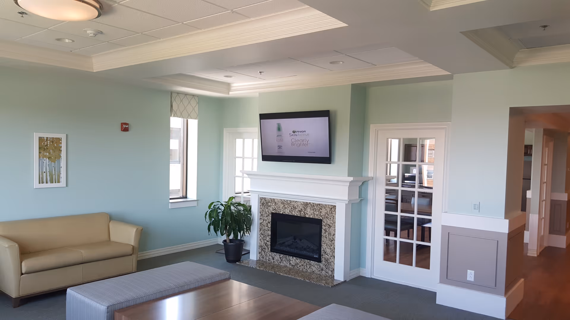 A bright and clean living room area with a beige sofa, a rectangular ottoman, a potted plant near a window, a fireplace with a granite surround, and a flat-screen TV mounted above the fireplace. The walls are painted light blue, and there is a framed artwork on one wall. French doors lead to another room with wooden flooring.
