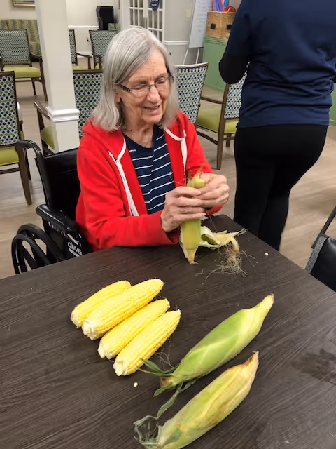 An elderly woman in a wheelchair wearing a red jacket and striped shirt is sitting at a table peeling corn. Several ears of corn, some husked and some still in their husks, are on the table. Another person is standing nearby with their back to the camera. The setting appears to be a communal indoor area with chairs and tables.