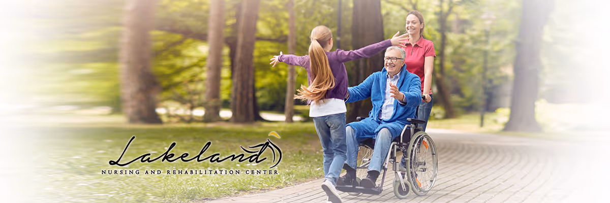 An elderly man in a wheelchair is being pushed by a smiling caregiver along a paved path in a park-like setting with trees. A young girl with long hair is running towards the man with open arms, suggesting a joyful reunion.