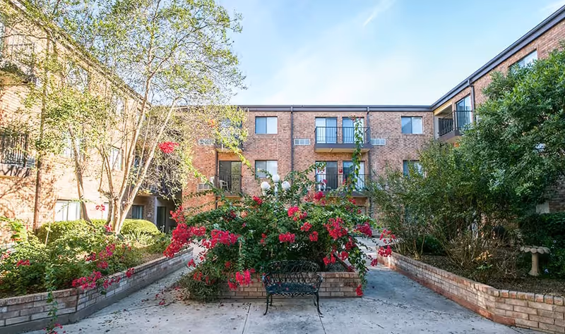 Outdoor courtyard area of a senior living facility with brick buildings surrounding it. The courtyard features a raised flower bed with vibrant red flowers and greenery, a black metal bench in front, and trees and shrubs along the sides. The sky is clear and blue.