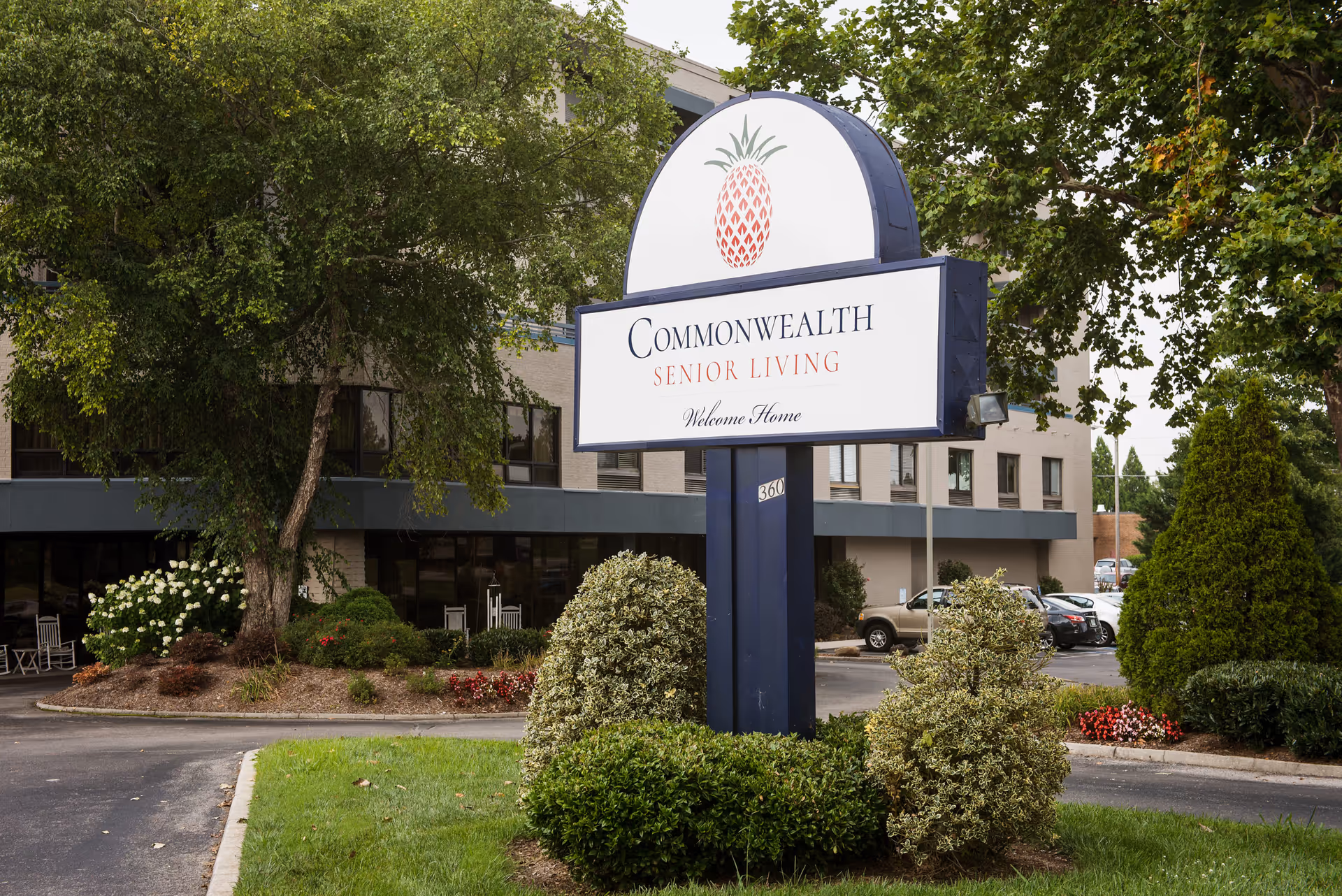 Outdoor view of the Commonwealth Senior Living facility sign with a pineapple logo, surrounded by bushes and trees, with the building and parking lot in the background.