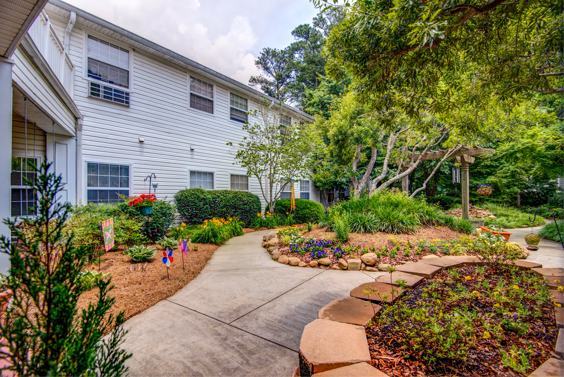 A landscaped garden area with a concrete pathway winding through colorful flower beds and green shrubs, adjacent to a white two-story building with multiple windows. Trees and a wooden pergola are visible in the background under a partly cloudy sky.