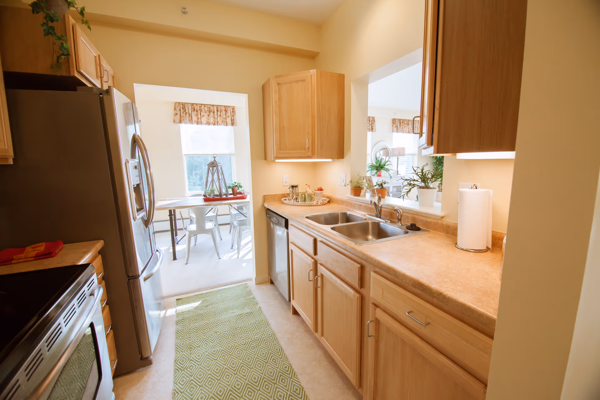 Bright kitchen with wooden cabinets, a double sink, stainless steel refrigerator and dishwasher, and a green patterned rug on the floor. Through an open doorway, a dining area with a table and metal chairs is visible, along with windows letting in natural light.
