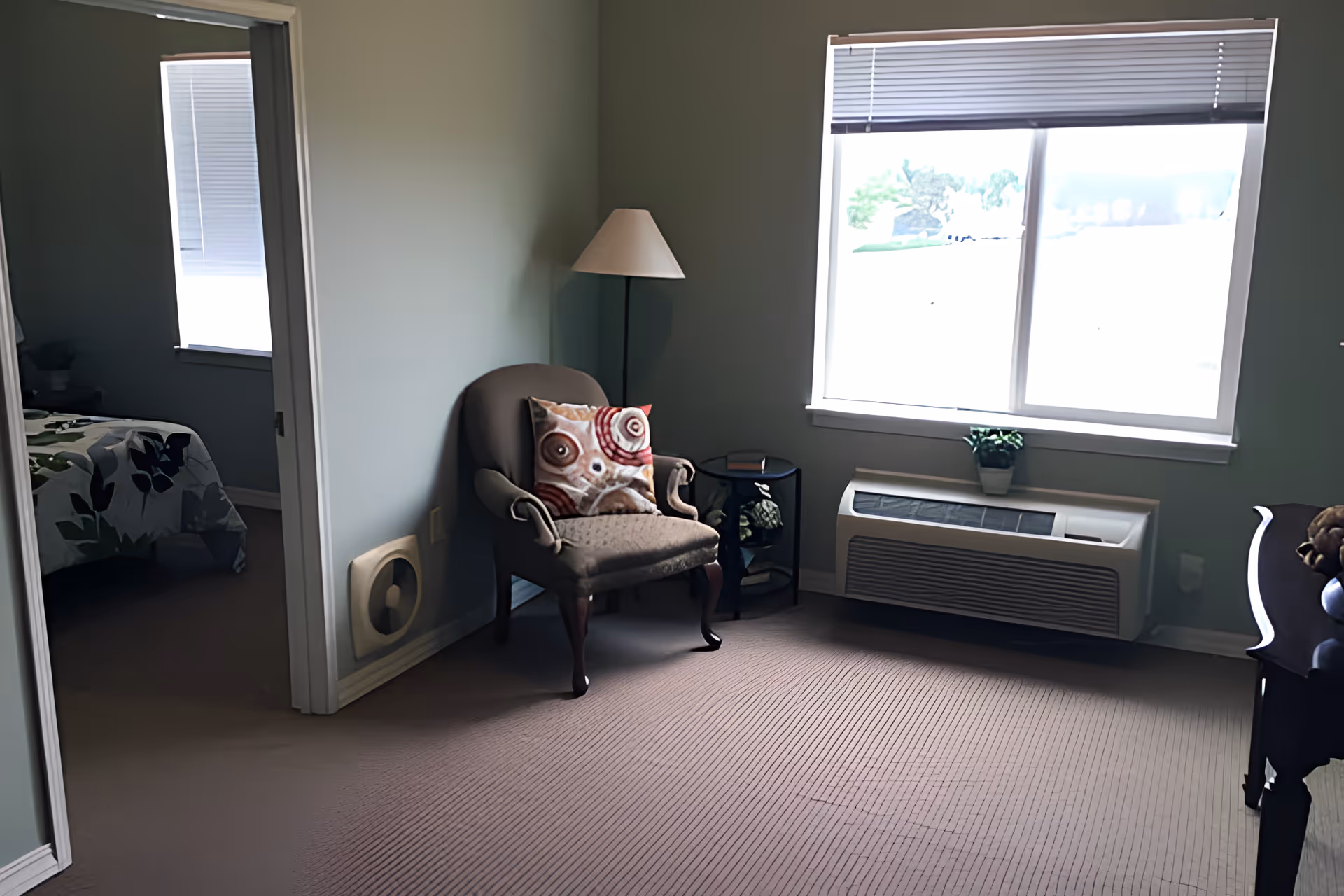 A cozy sitting area with a cushioned armchair featuring a decorative pillow, a floor lamp, a small side table with a book, and a window with blinds partially open letting in natural light. Adjacent to the sitting area is a bedroom with a bed covered in a floral-patterned bedspread.