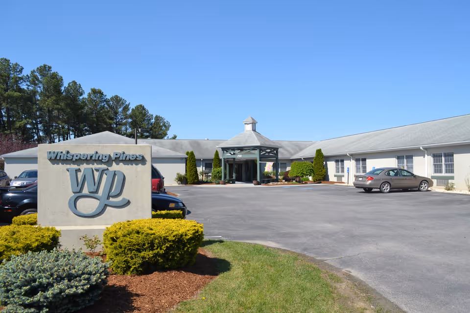 Exterior front view of Whispering Pines assisted living showing a stone WP sign, driveway and parking lot, and the building's entrance canopy.