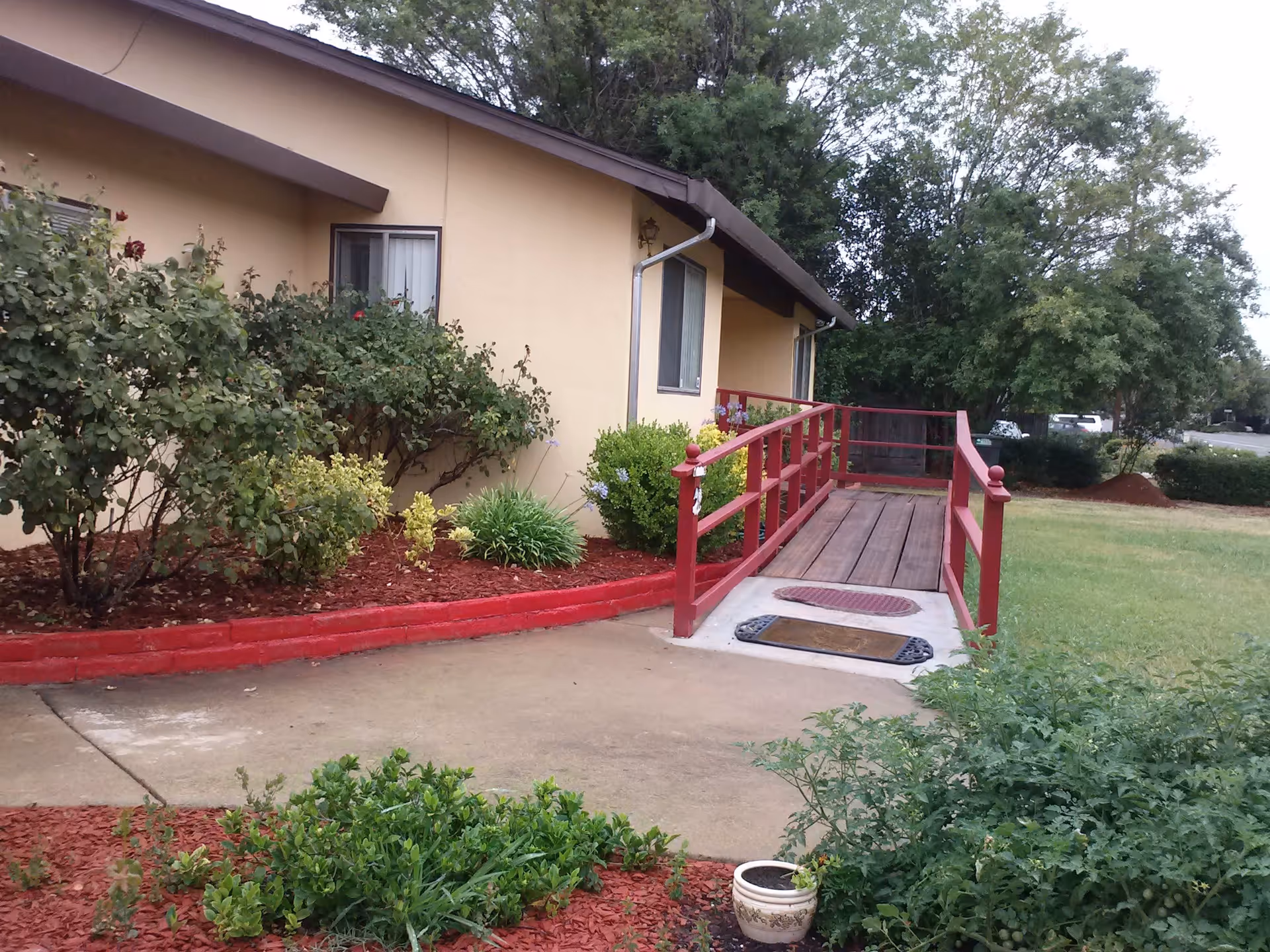 Exterior view of a single-story building with beige walls and a brown roof, featuring a wooden ramp with red railings leading to the entrance. The surrounding area has green grass, bushes, and trees, with a concrete walkway and red mulch landscaping.