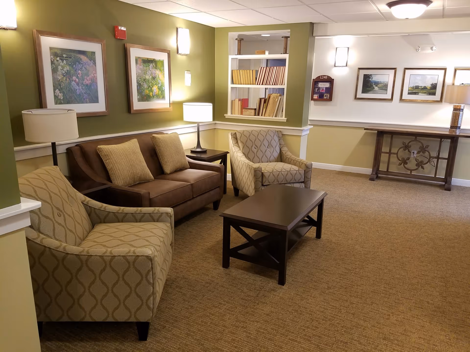 A cozy sitting area in a senior living facility with a brown sofa, two patterned armchairs, a dark wooden coffee table, and two side tables with lamps. The walls are painted green and white, adorned with framed floral and landscape artwork. There is a built-in bookshelf with books and a decorative console table against the far wall.