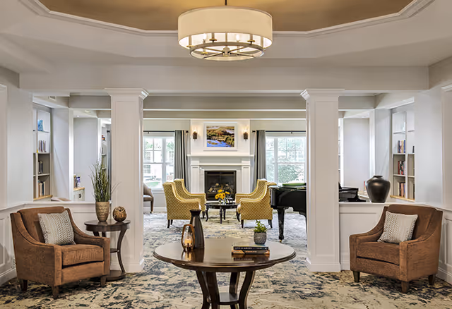 A bright and elegant senior living common area featuring two brown armchairs with pillows on either side of a round wooden table with decorative items. The room has white columns and built-in shelves with books and decor. In the background, there are yellow armchairs arranged around a fireplace with a painting above it, and large windows letting in natural light. A grand piano is also visible near the fireplace.