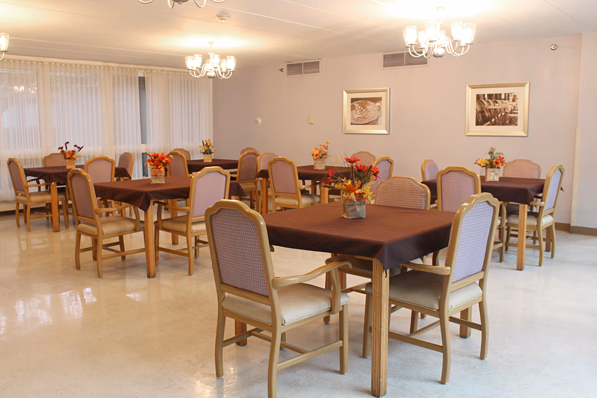 A dining room with several wooden tables covered with dark tablecloths, each surrounded by matching wooden chairs with cushioned seats and backs. Each table has a small floral centerpiece. The room has light-colored walls, two framed pictures, large windows with sheer curtains, and two chandeliers hanging from the ceiling.