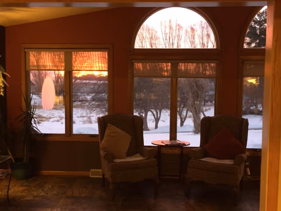A cozy interior seating area with two cushioned armchairs, each with a pillow, positioned on either side of a small round wooden table. Behind the chairs are large windows showing a snowy outdoor scene with trees and a sunset sky.