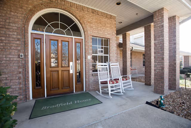 Front entrance of a brick building with a wooden door featuring decorative glass panels and an arched window above. Two white rocking chairs with red and white striped cushions sit on the covered porch next to a green doormat that reads 'Bradfield House'. The porch is supported by brick columns and there is landscaping with rocks and a small garden gnome nearby.