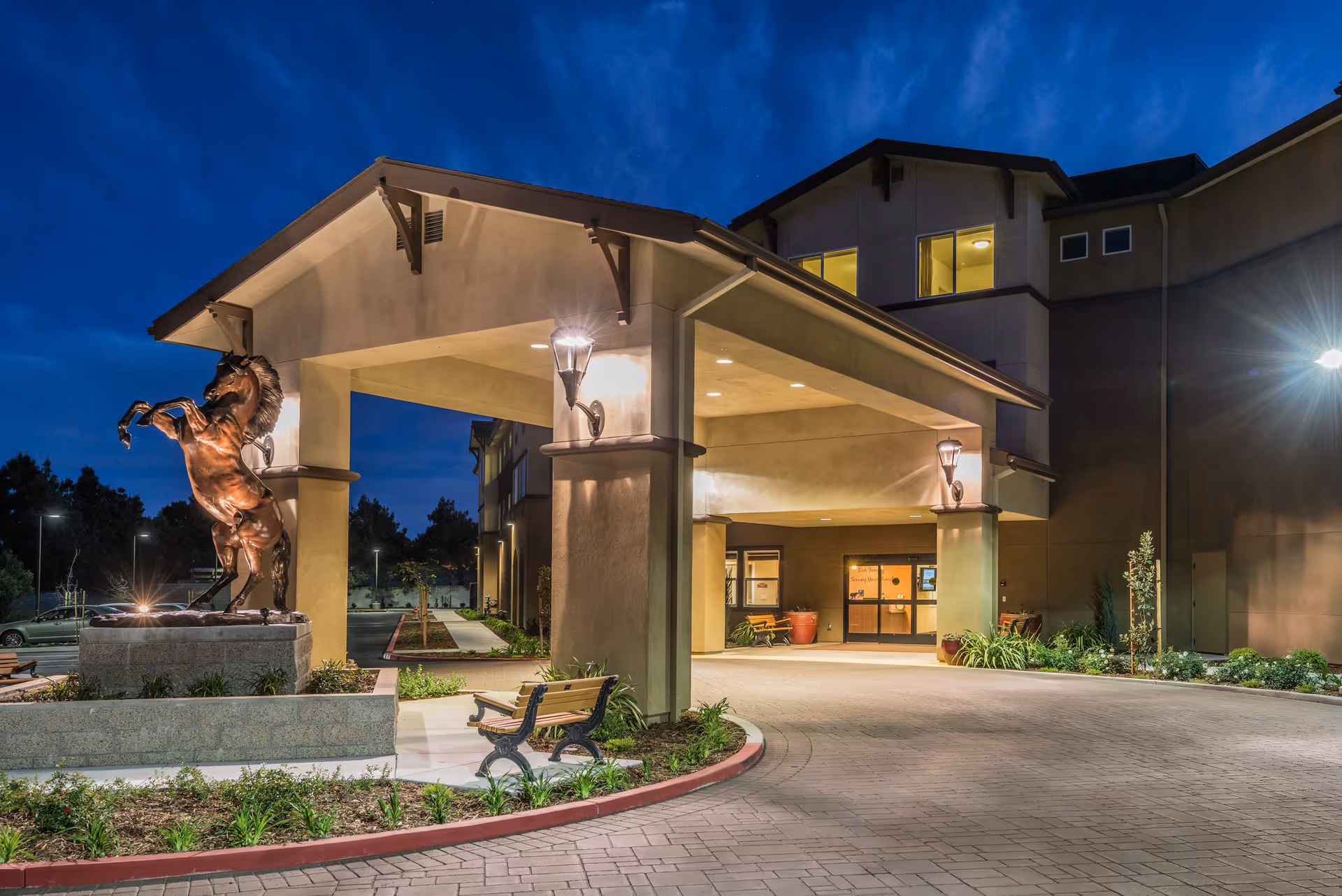 Entrance of Carlton Senior Living Davis at dusk with a covered drop-off area, a bronze statue of a rearing horse on a pedestal, benches, and well-maintained landscaping.