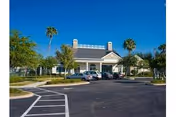 Exterior view of a single-story building with a pitched roof, surrounded by palm trees and a parking lot with several parked cars under a clear blue sky.