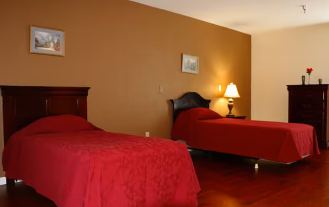 Two twin beds with red bedspreads, a nightstand with a lamp, framed artwork, and a dresser in a furnished bedroom.