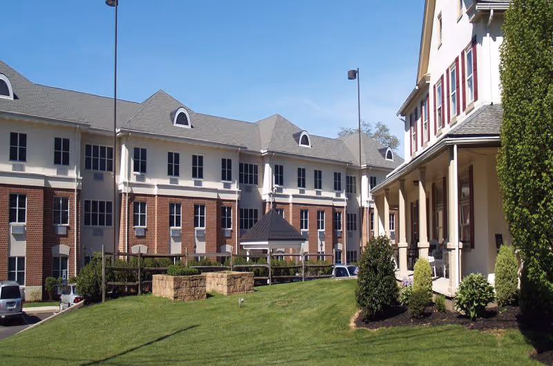 Front exterior of a multi-story red-brick and cream senior living building with a lawn, porch, and clear blue sky.