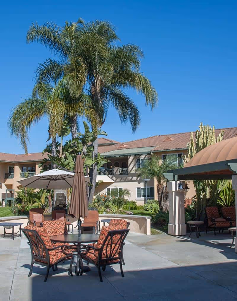 Outdoor patio area with cushioned chairs around a round table, umbrellas, palm trees, and a building with balconies in the background under a clear blue sky.
