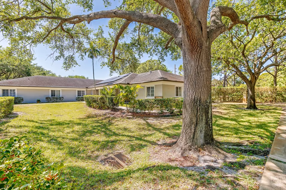 Exterior view of a single-story building surrounded by green grass, trees, and shrubs under a clear blue sky at Cottages at Palm Beach.