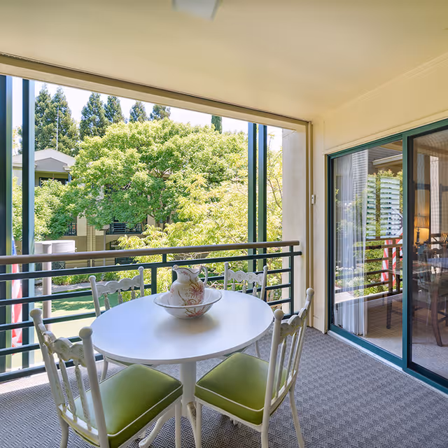A covered balcony with a round white table and four white chairs with green cushions. On the table is a decorative pitcher and bowl. The balcony overlooks green trees and parts of the building exterior. Sliding glass doors lead inside to a room with a lamp and furniture visible.