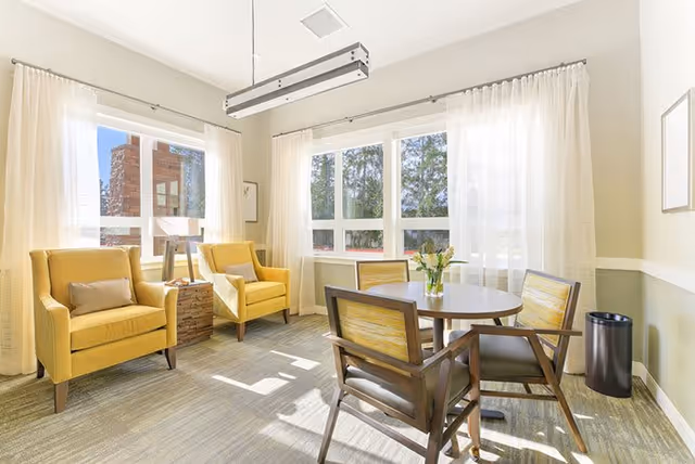 A bright and airy room with large windows covered by sheer white curtains. The room features two yellow armchairs with a small wooden side table between them, and a round wooden table surrounded by four wooden chairs with yellow cushions. A vase with flowers is placed on the table. The walls are light-colored, and the floor is carpeted. A modern rectangular ceiling light fixture hangs above the table.