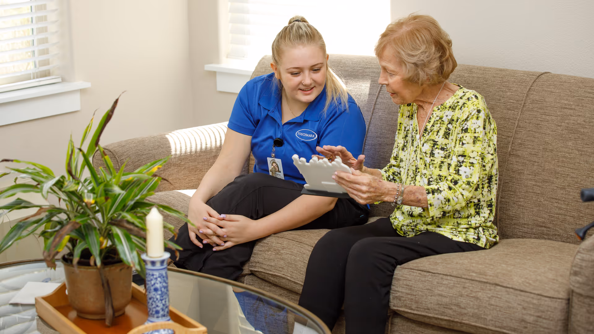 A young female caregiver in a blue Touchmark uniform sits on a beige couch next to an elderly woman wearing a yellow patterned blouse. They are looking at and discussing a white object the elderly woman is holding. A glass coffee table with a potted plant and a candle is in front of them, and sunlight streams through the window blinds in the background.