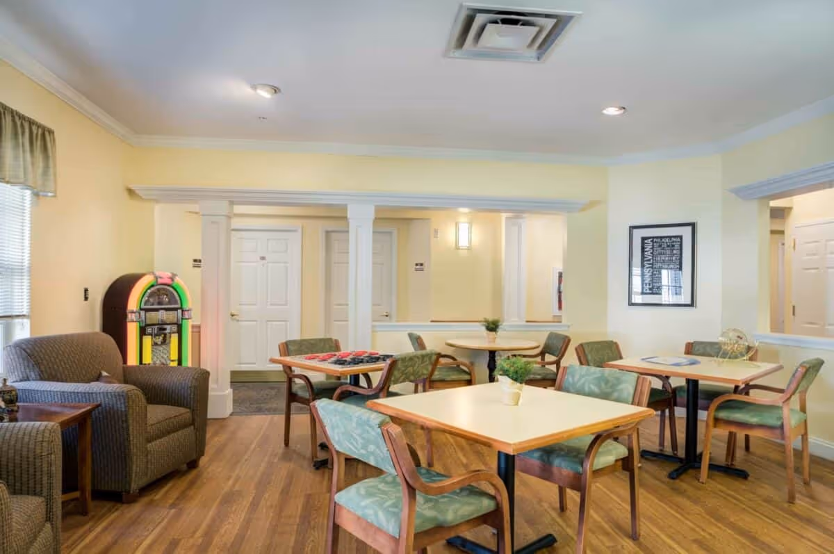 A cozy common area with several tables and chairs arranged for socializing or games. There are green cushioned chairs around light-colored wooden tables, a couple of armchairs, and a colorful jukebox against a yellow wall. The room has wooden flooring, soft yellow walls, and a framed picture on the wall.
