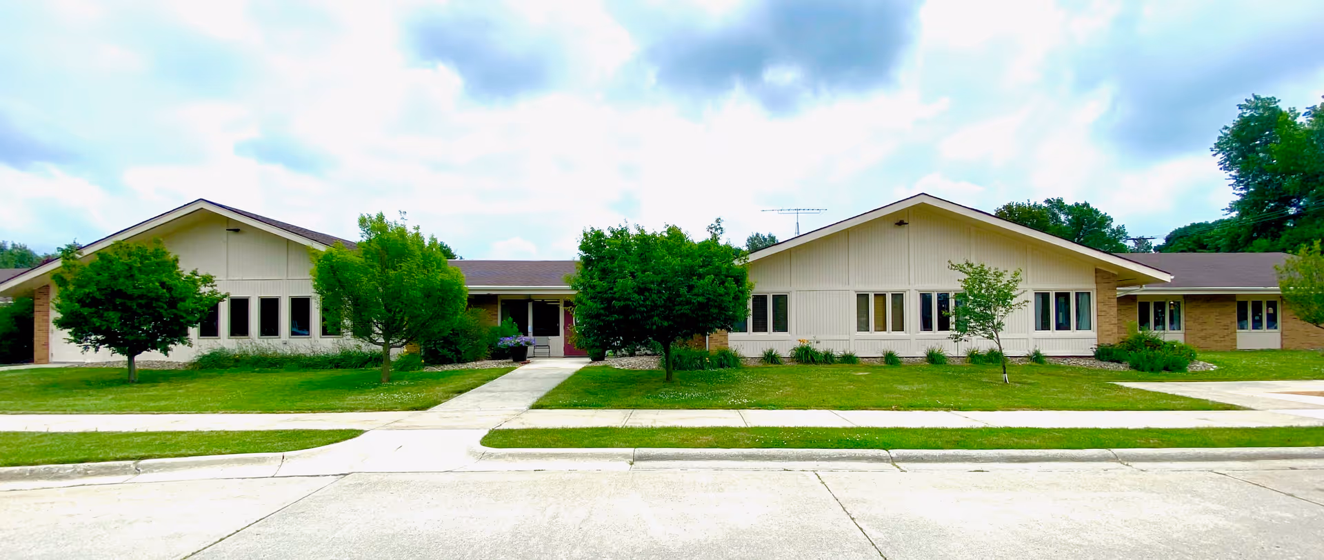 Front exterior of a single-story senior living facility with a lawn, trees, and sidewalks under a cloudy sky.