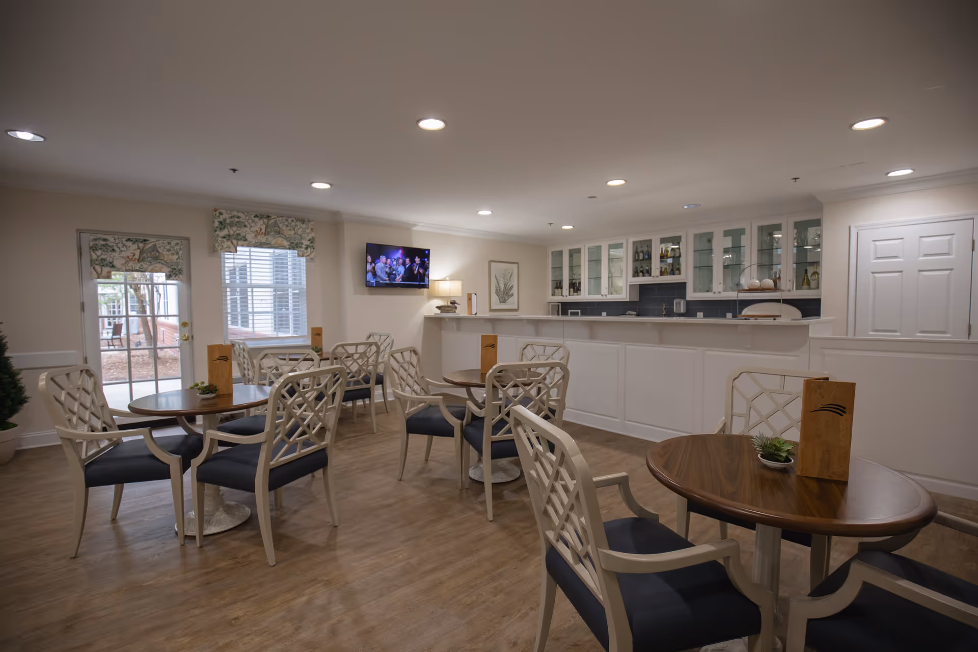 Communal dining/lounge area with round wooden tables and lattice-back chairs facing a white serving counter and wall-mounted TV.
