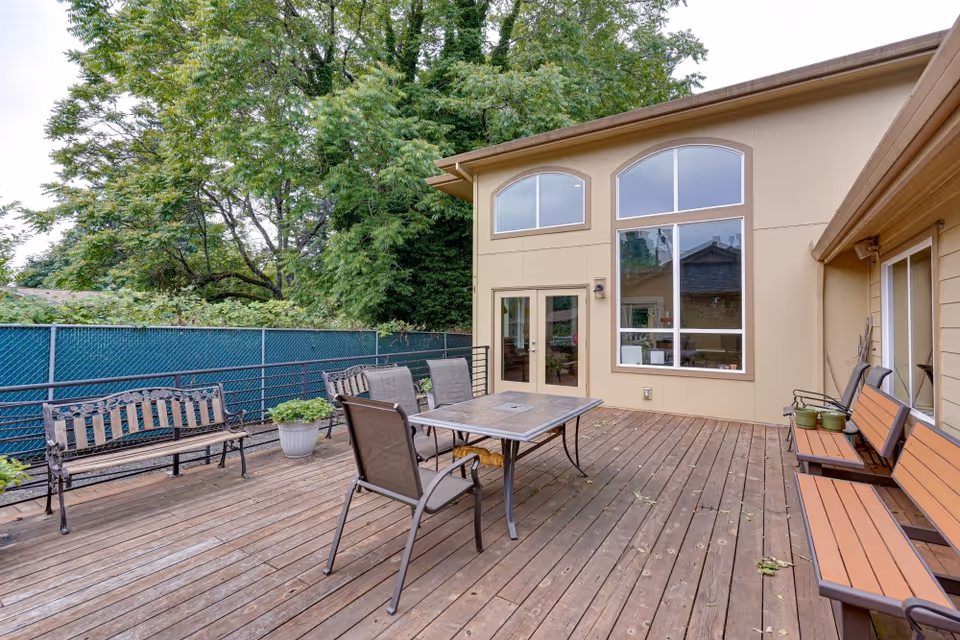 Outdoor wooden deck area with metal and wood benches, a table with four chairs, potted plants, and a beige building with large windows and a door. Trees and a green fence are visible in the background.