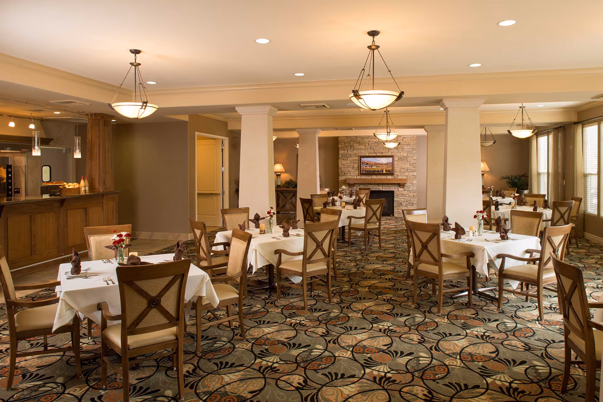 A spacious dining room with multiple tables covered in white tablecloths, each set with brown napkins, cutlery, and small flower vases. The room features patterned carpet flooring, beige walls, and several hanging light fixtures. There is a stone fireplace with a framed picture above it in the background, and large windows with curtains on the right side. A wooden counter area is visible on the left side.
