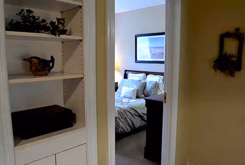 View through an open doorway into a bedroom with a neatly made bed featuring multiple pillows. To the left of the doorway is a built-in white shelving unit with decorative items including a small basket and a figurine. The walls are painted in neutral tones, and a framed picture hangs above the bed.