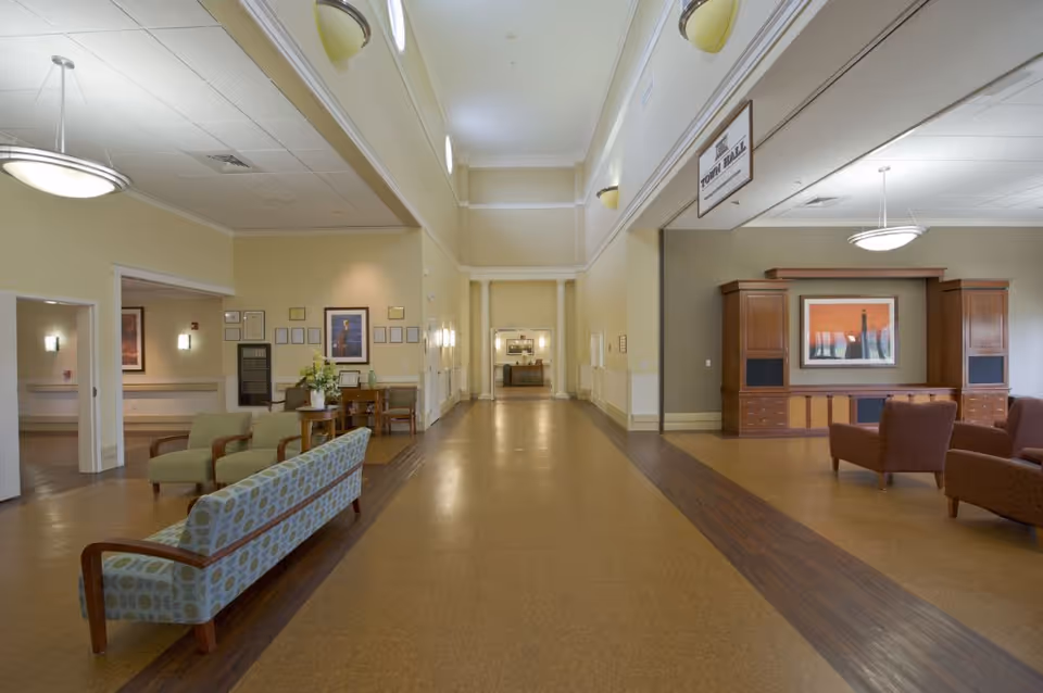 A spacious, well-lit interior hallway or common area in a senior living facility with high ceilings and circular windows near the ceiling. The area features seating arrangements including patterned sofas and armchairs, a wooden entertainment center with a framed artwork above it, and a sign hanging from the ceiling labeled 'Town Hall'. The walls are painted light yellow and beige, with framed pictures and wall sconces providing additional lighting.