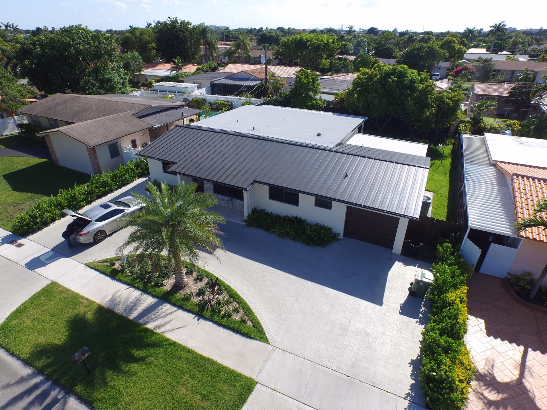Aerial view of a single-story residential building with a dark metal roof, a driveway with a silver car parked and a person standing near the car's open trunk. The property has a well-maintained lawn with a palm tree and surrounding greenery. Neighboring houses and trees are visible in the background under a clear blue sky.