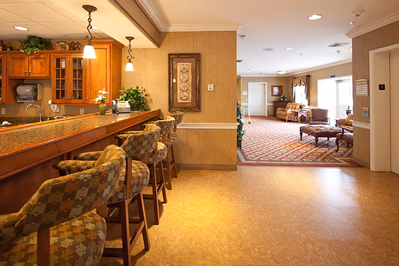 Interior view of a senior living facility showing a bar area with patterned cushioned bar stools and wooden cabinetry on the left, and a lounge area with sofas, armchairs, and a patterned carpet in the background. The walls are beige with framed artwork, and there is an elevator on the right side.
