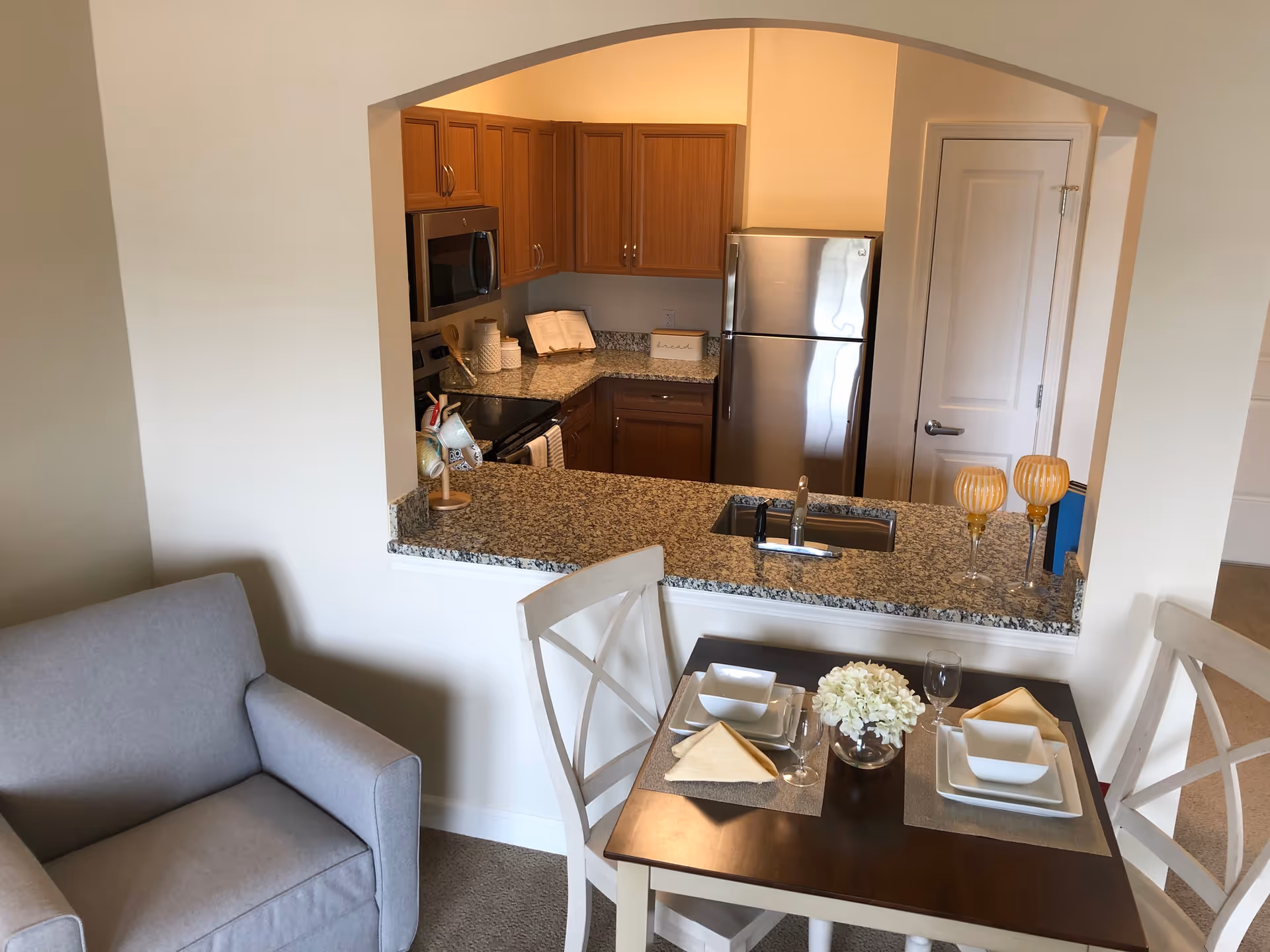 View of a small kitchen with wooden cabinets, a stainless steel refrigerator, microwave, and stove. In front of the kitchen is a granite countertop with a sink and two decorative candle holders. Adjacent to the kitchen is a dining table set for two with white plates, bowls, wine glasses, beige napkins, and a small flower arrangement. A gray upholstered armchair is positioned next to the dining area.