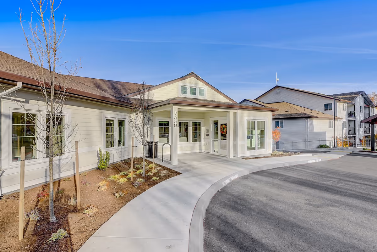 Exterior view of a senior living facility named Courtyard at Mt. Tabor showing the entrance area with a covered drop-off, several windows, young trees planted along the sidewalk, and a clear blue sky.