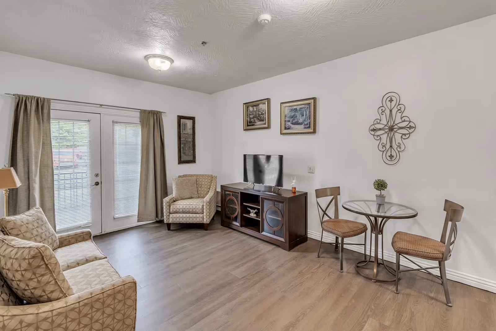 Bright living room with armchairs and a sofa, a TV on a console, a small round dining table with two chairs, and glass doors with curtains.