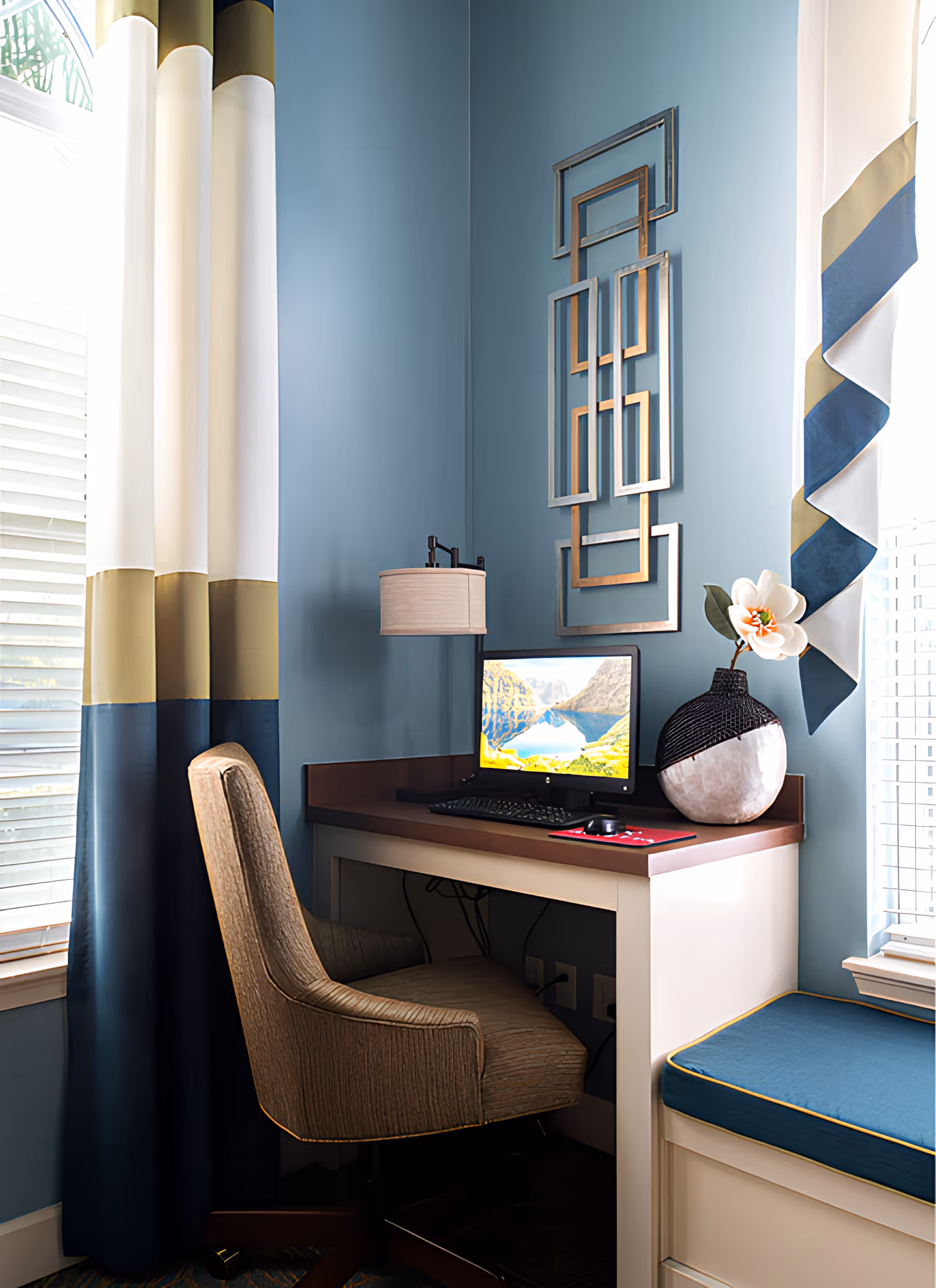 A cozy corner workspace with a beige upholstered chair, a wooden desk with a computer monitor displaying a scenic mountain and lake wallpaper, a decorative vase with a white flower, and geometric wall art on a blue wall. The space is flanked by windows with blue, white, and gold curtains and a cushioned window seat with blue upholstery and yellow piping.