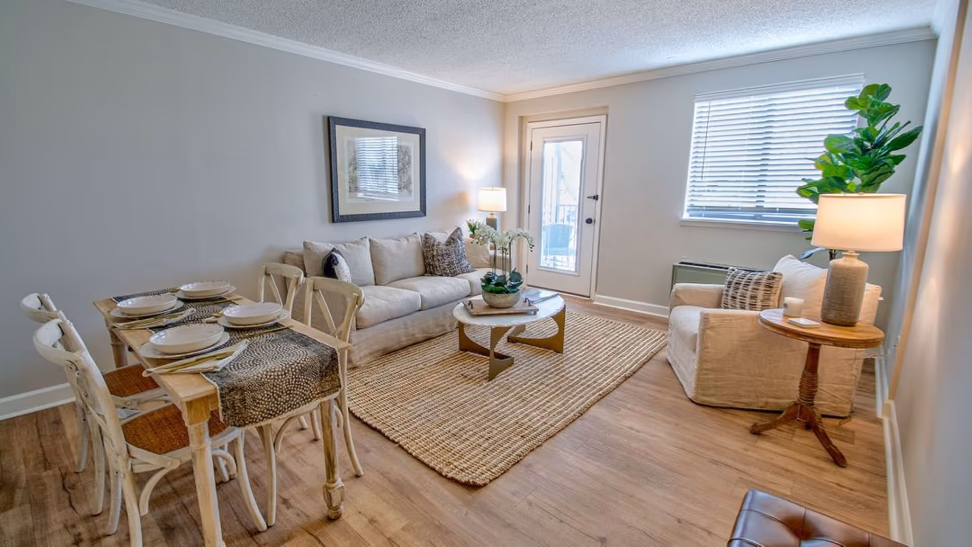 Bright furnished living room with a sofa, armchair, coffee table, dining table set, and a door to a balcony.