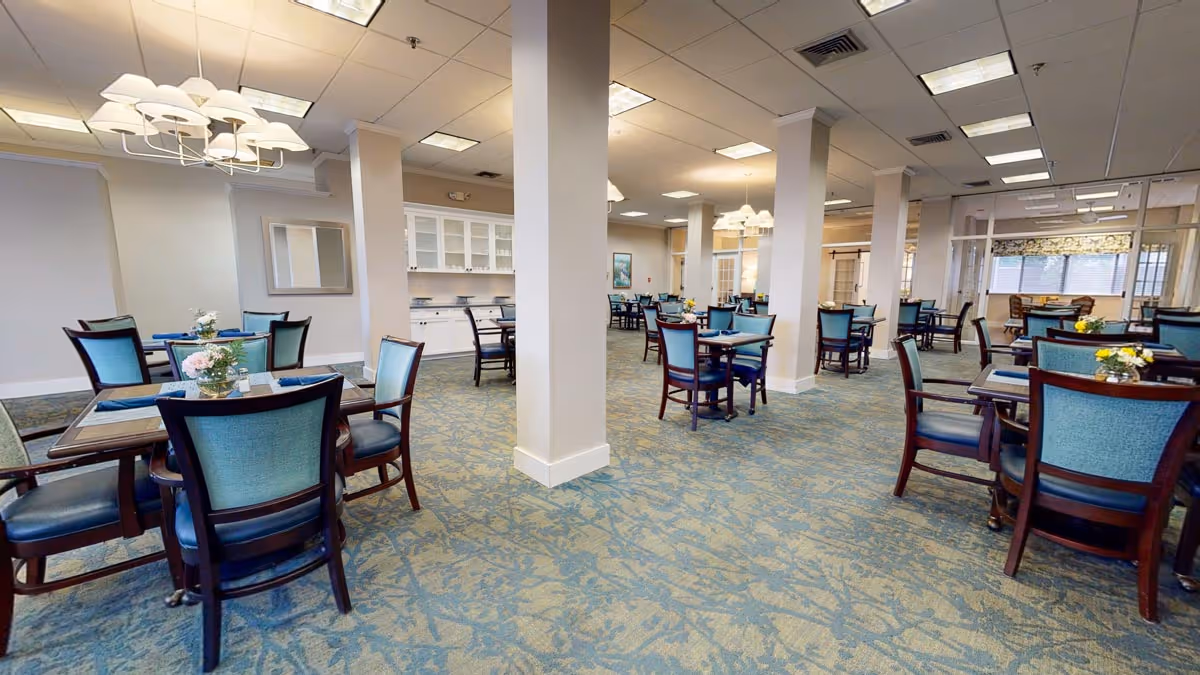 Spacious dining room with multiple tables and teal-upholstered chairs on patterned carpet under ceiling lights.