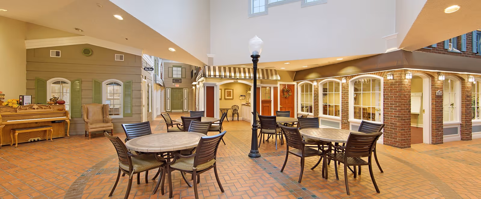 Indoor common area designed to resemble an outdoor street scene with round tables and chairs, a street lamp, a piano, and building facades with windows and doors under a high ceiling with windows.