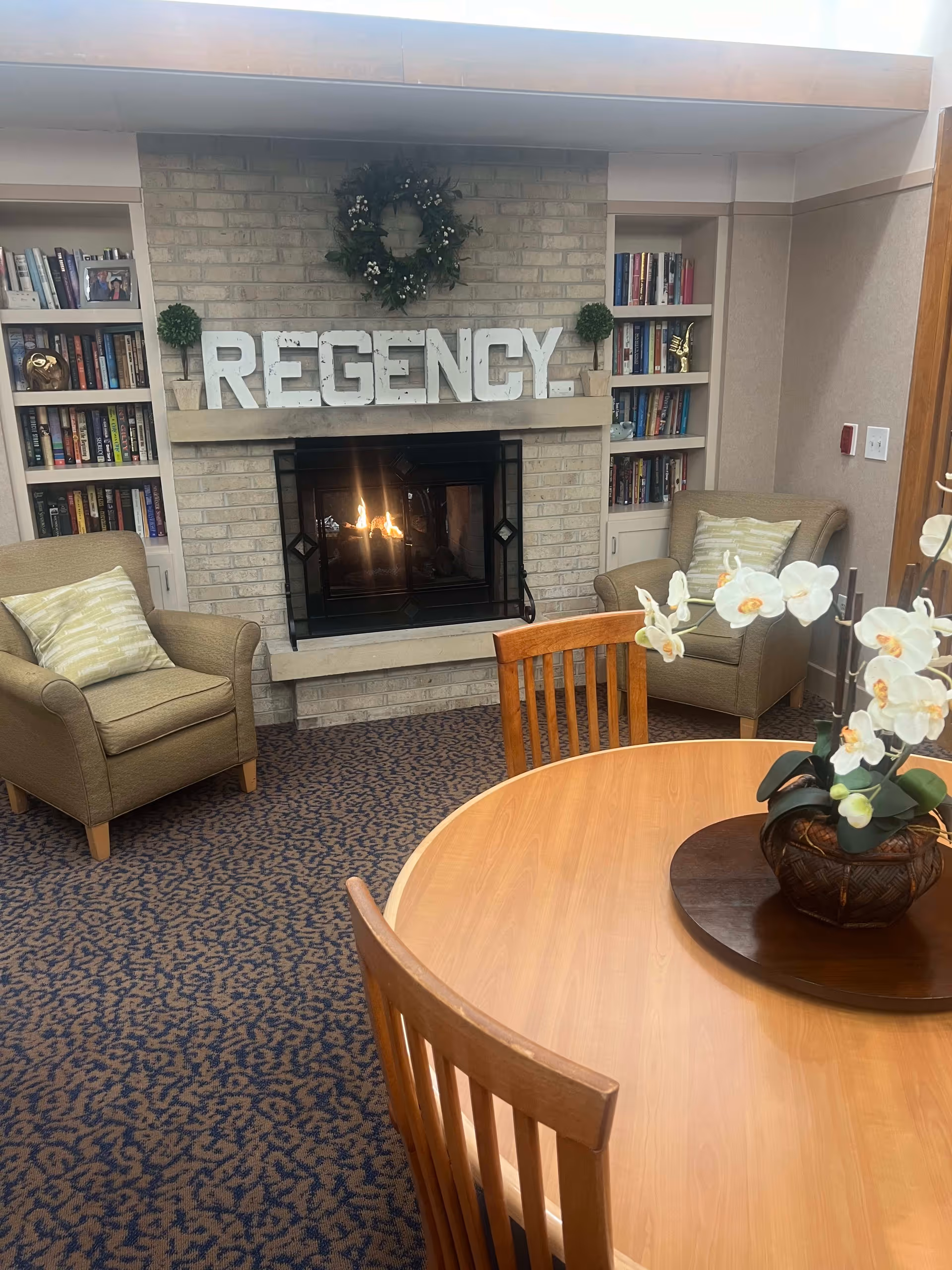 Cozy senior living community common area with a lit fireplace set in a brick wall, flanked by built-in bookshelves filled with books and decorative items. Two cushioned armchairs with striped pillows are placed on either side of the fireplace. In the foreground, there is a round wooden table with wooden chairs and a potted white orchid centerpiece.