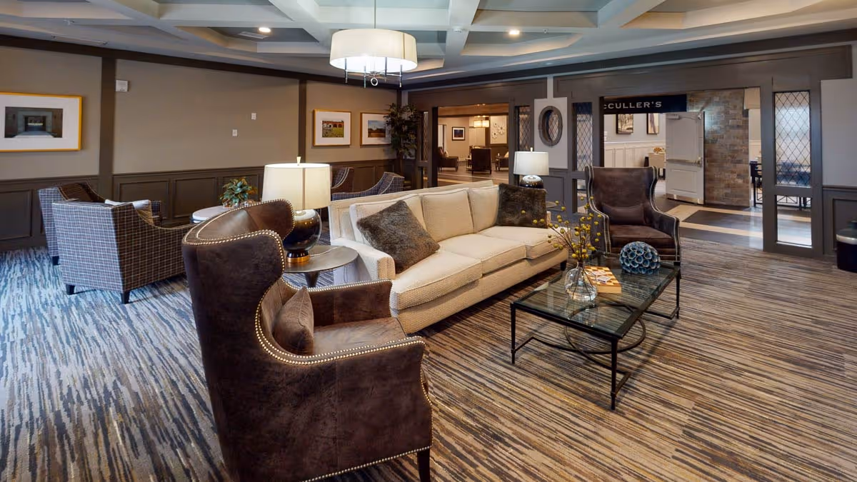 A cozy living room area in a senior living facility featuring a beige sofa with dark brown pillows, two dark brown armchairs with nailhead trim, a glass coffee table with decorative items, and a side table with a lamp. The room has patterned carpet flooring, framed artwork on the walls, and a ceiling light fixture. In the background, there is an open doorway leading to another room.