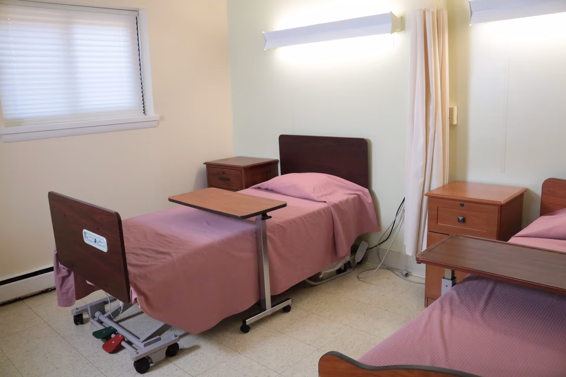 A simple room with two hospital-style beds covered in pink sheets, each with an overbed table and a wooden nightstand beside it. The room has a window with closed blinds, a curtain divider, and light-colored walls and floor.