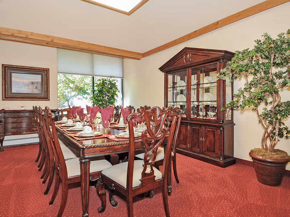 A formal dining room with a long wooden dining table set for a meal, featuring folded burgundy napkins, white plates, and cups. The room has a red carpet, a large wooden china cabinet displaying plates and glassware, a large potted plant, and a window with a view of greenery outside. There is also a framed painting on the wall and a wooden sideboard.