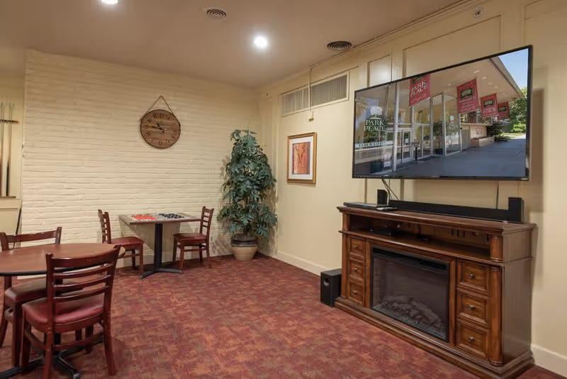 A cozy interior room with red carpet and cream-colored walls. There are two small tables with chairs, one table has a checkers game set up. A large flat-screen TV is mounted on the wall above a wooden cabinet with an electric fireplace. A potted plant and a wall clock are also visible.