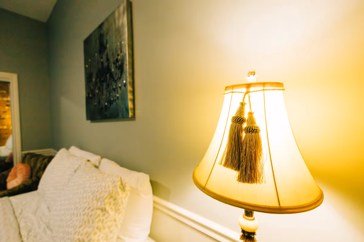 A cozy living room area featuring a lit table lamp with decorative tassels, a white couch with patterned pillows, and a piece of wall art hanging above the couch.
