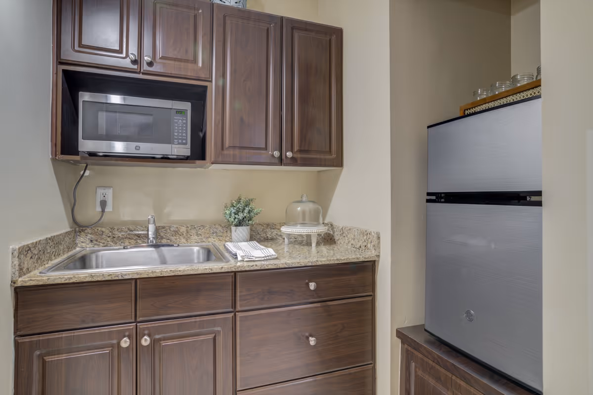 A small kitchen area with dark wood cabinets, a stainless steel microwave mounted above a granite countertop with a sink, a small potted plant, a folded towel, and a glass cake stand. To the right, there is a stainless steel refrigerator.