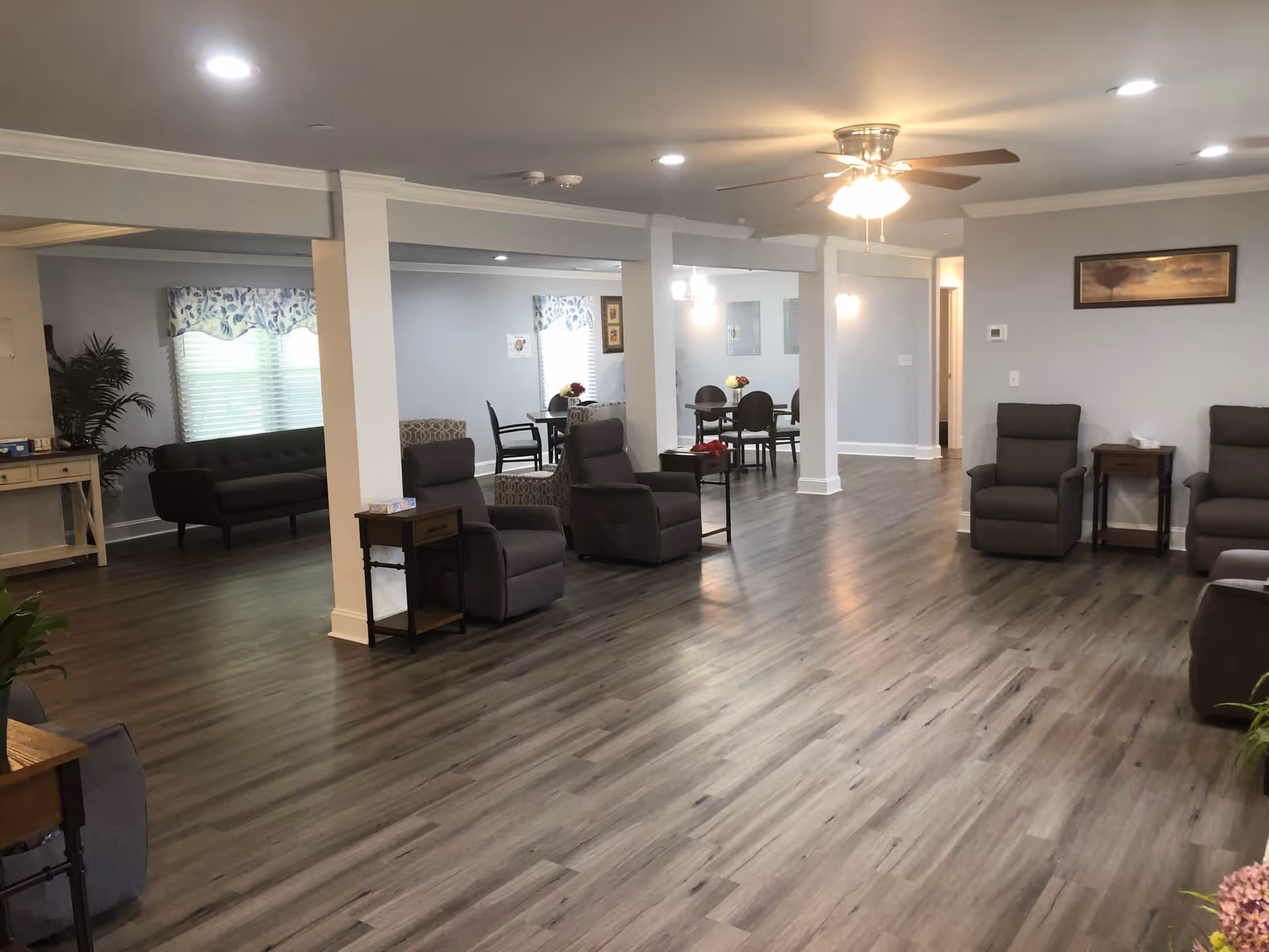 Spacious senior living common area with multiple recliner chairs, sofas, a dining table, ceiling fan, and wood‑look flooring.