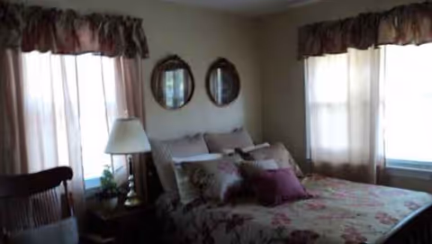 Bedroom with floral bedspread, pillows, and curtained windows in a senior care home.